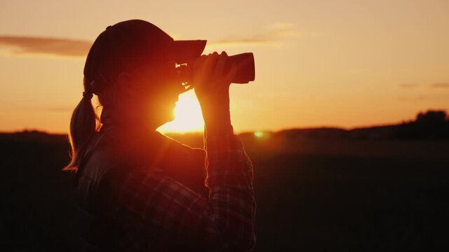 Silhouette of woman in baseball cap using binoculars at sunset in a field.