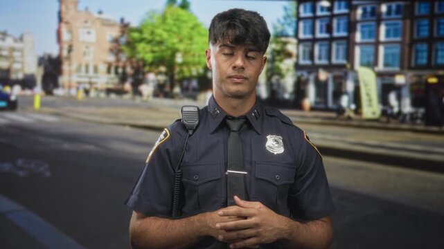 Young hispanic police officer wearing navy uniform and tie, badge visible, radio mic and hands clasped while making hand gestures on a busy street; duty concern.
