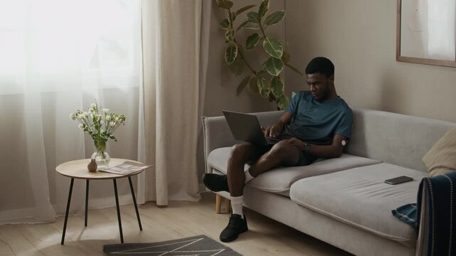 Wide angle shot of young adult African American man with diabetes typing on laptop while sitting on couch in modern living room decorated with green plants, slow motion