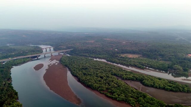Cinematic aerial view of Talpona River flowing through dense mangrove forests, with exposed sandbanks and bridges stretching across the water beneath a vast hazy sky.