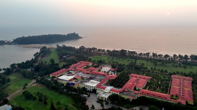 Cinematic aerial view of a luxury beachfront resort in Goa featuring expansive red-roofed architecture, landscaped gardens, and palm-lined open spaces facing the calm Arabian Sea at sunset.