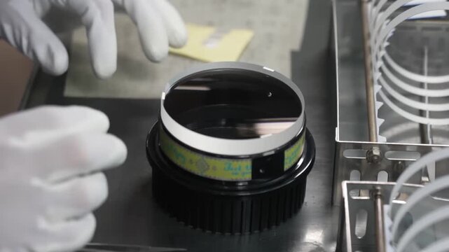 Gloved hands carefully inspecting large diameter coated optical lens element with metal housing in cleanroom