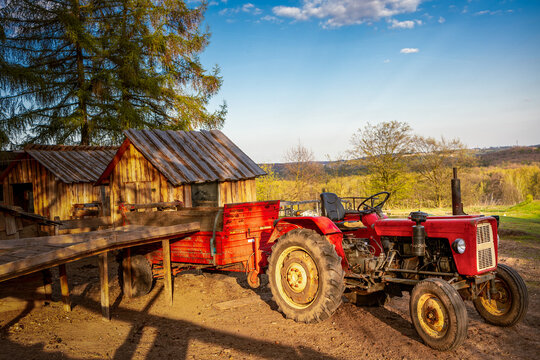 Old vintage red tractor with a cargo trailer parked next to rustic wooden sheds on a local farm.A high-quality rural scene captured during warm evening golden hour, perfect for organic farming, small 