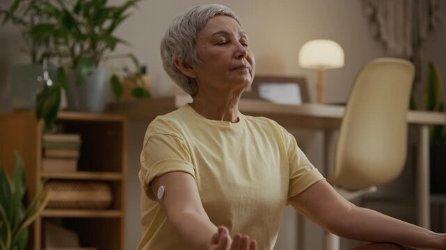 Tilt down shot of senior woman wearing wearable glucose monitoring device on upper arm relaxing in lotus pose on mat during home meditation session
