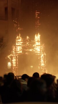 Falla figure atop burning wooden frame during Las Fallas crema in Valencia, Spain, crowd silhouettes below and building facade lit orange by flame