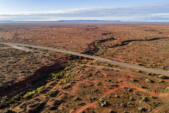Aerial view of a long straight road crossing a bridge over a dry red creek bed in the vast outback landscape under a clear sky Flinders Ranges, South Australia, Australia.