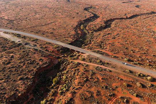 Aerial view of a long asphalt road and bridge crossing a winding dry creek bed in the vast outback landscape with a lone vehicle in Flinders Ranges, South Australia, Australia.
