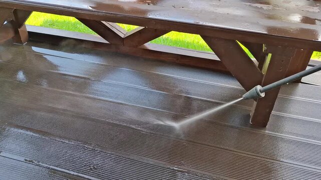 High pressure washer cleans a wet wooden deck and patio furniture outdoors, showing home maintenance, restoration and exterior cleaning service.