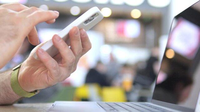 Hand holds smartphone, taps screen. Other hand types on laptop. Background blurred, indoor setting. Lights glow softly. Person multitasks with devices. Focus on hands and tech interaction
