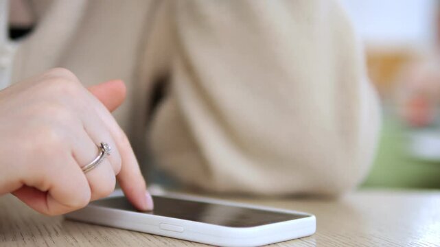 Hand with a ring taps a white smartphone on a wooden table. The person wears a beige sweater. Background is softly blurred with warm tones. Focus remains on the hand and device interaction