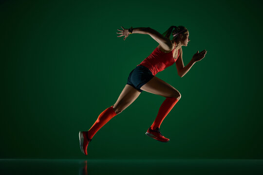 Focused woman runner sprinting forward on dark green studio floor. Concept of speed workout, sports performance, running apparel, fitness ads and active lifestyle branding.