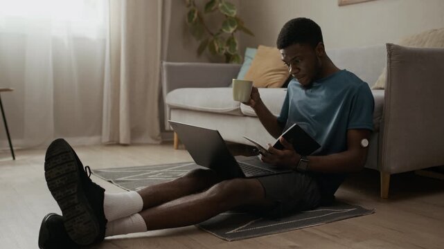 Low eye level shot of relaxed young Black man with diabetes drinking tea while planning workday with laptop and notebook sitting on living room floor while CGM tracking blood sugar level, slow motion