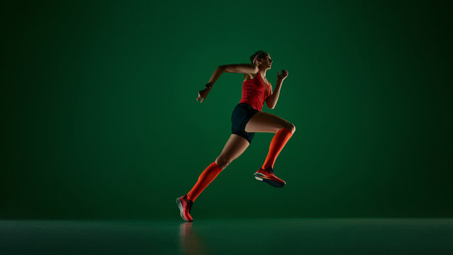 Side view of female runner accelerating on green studio background. Concept of running technique, speed training, sport gear promo, fitness coaching and endurance lifestyle.