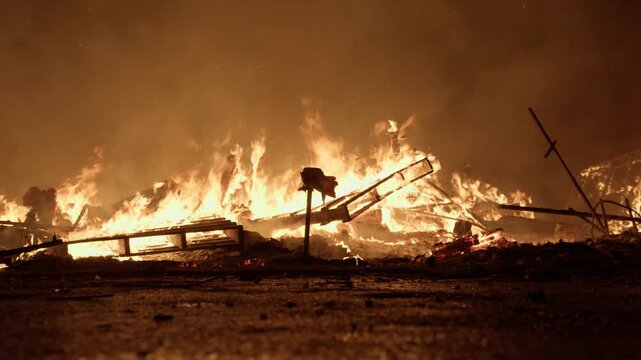 Low angle view of falla wreckage burning on wet pavement during Las Fallas crema in Valencia, Spain, scattered debris and metal frame fragments glowing
