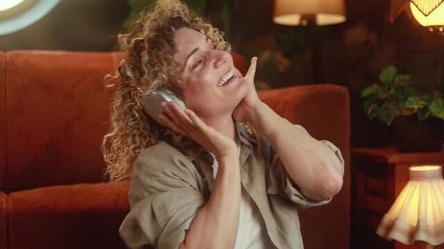 Happy curly haired woman listening to music in headphones and enjoying relaxing evening at home with positive emotions. High quality 4k footage