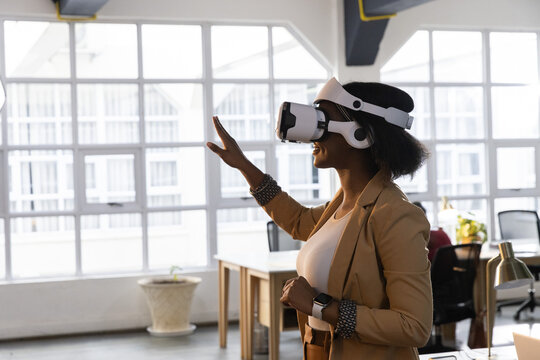 African American woman wearing VR, tan blazer reaching interface near office windows, copy space