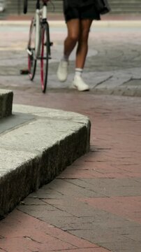 Vertical video: Approaching, rider pushing red-rim bike across plaza, walker crossing with tan bag