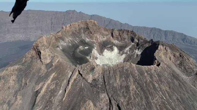 Aerial close-up of the volcanic crater at Pico do Fogo, Cabo Verde. The footage highlights the rugged basaltic rim, steep slopes, and ash-covered geological formations of the summit