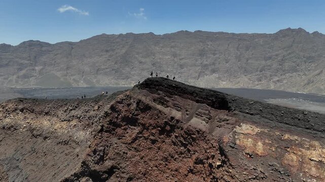 Drone shot of hikers trekking on the rugged crater rim of Pico do Fogo, Cabo Verde, with the dramatic Bordeira wall and volcanic plains of Ch&atilde; das Caldeiras in the background
