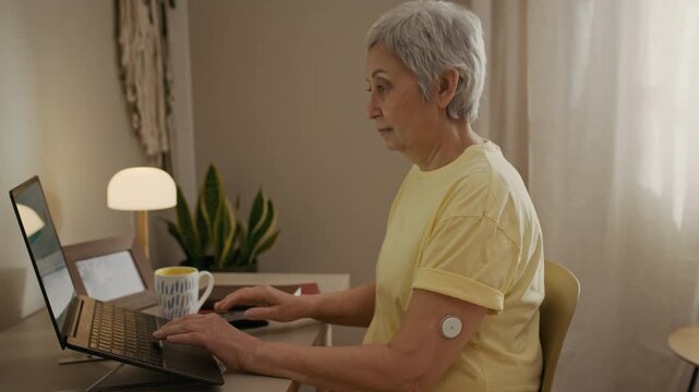 Side view shot of senior woman typing on laptop studying in online course from home while CGM sensor on upper arm tracking glucose levels, daily diabetes management concept