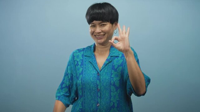 Woman smiles and makes ok hand gesture with bare right hand, wearing blue floral shirt and hoop earring against soft blue studio backdrop; approval joy confidence.