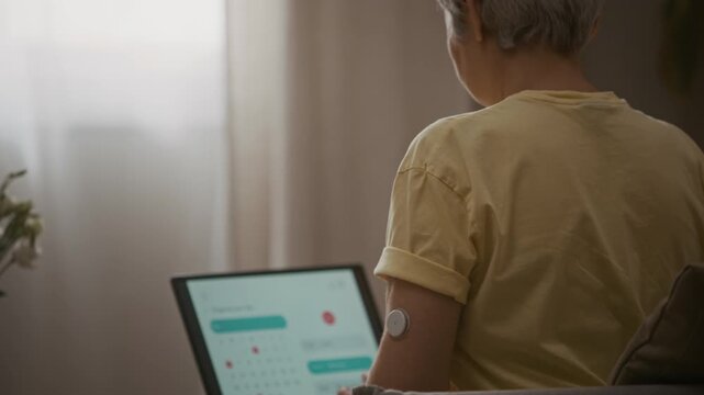 Rear view close up shot of senior woman wearing CGM diabetes sensor on upper arm managing daily tasks on laptop at home while device monitoring blood sugar level, copy space