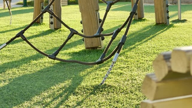 A sturdy climbing rope bridge hangs low over a vibrant green lawn in a quiet playground. This peaceful scene captures the simple joy and adventurous spirit of a sunny day outdoors.