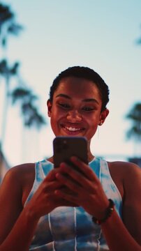  joyful young Black woman stands in evening city with by tall palm trees, laughing while looking at smartphone. Female exudes happiness and genuine excitement, embodying joy of digital communication
