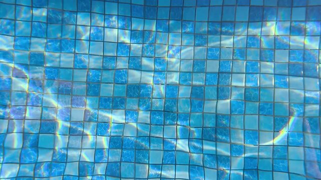 Close-up of rippling water in a swimming pool with blue mosaic tiles and sunlight reflections on the surface.