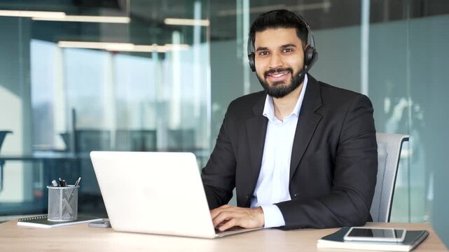Portrait of a happy businessman in a headset sitting at a desk at a workplace working on a computer in business office. Smiling male corporate operator in suit agent of call center looking at camera