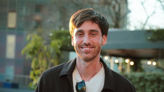 A cheerful young man stands outdoors in a casual outfit, exuding confidence and warmth. The golden hour sunlight highlights his features, creating a vibrant atmosphere in an urban environment.