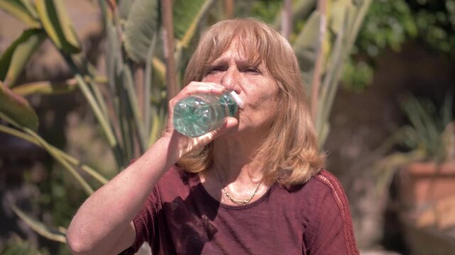 Mature blonde woman drinks water from a bottle in a sunny garden while trying to refresh herself and stay hydrated during an extremely hot summer day.