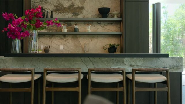 Modern kitchen shows marble counter and backsplash with natural veins, dark shelves holding jars and vases, tall glass vase with bright pink flowers adds lively contrast