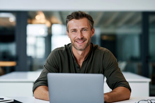 Smiling confident middle aged business man entrepreneur working on laptop in modern office. Happy professional executive manager looking at camera sitting at corporate desk using computer technology 