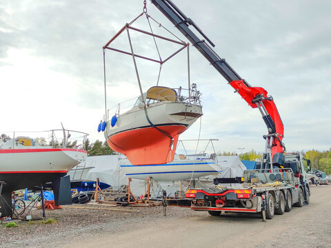 A heavy-duty truck with a large red crane prepares to lift a white sailboat out of its storage cradle in a marina yard under an overcast sky