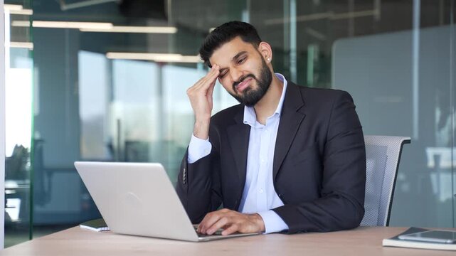 Unhealthy businessman suffering from headache while working on laptop sitting at workplace in office. Unwell worker holding and massaging his forehead and temples with his hands. He has a migraine