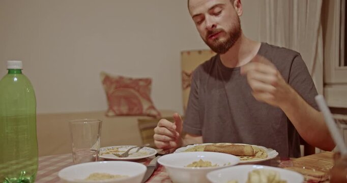 A young man with a beard enjoys a casual evening meal at home, picking food from a bowl and eating it. He appears relaxed and comfortable in the cozy dining setting.