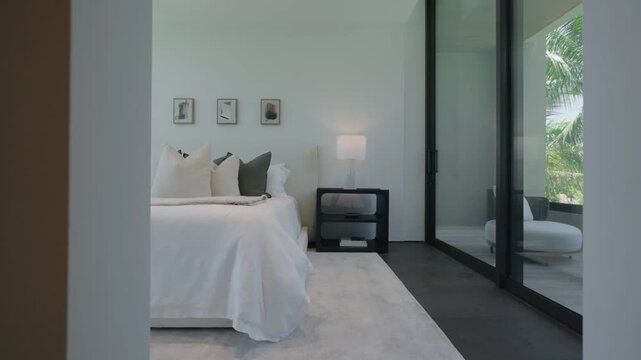 Modern bedroom shows white bed with dark pillows and bedside table, lamp softly lights clean space, doorway frames view creating calm and elegant atmosphere with simple design