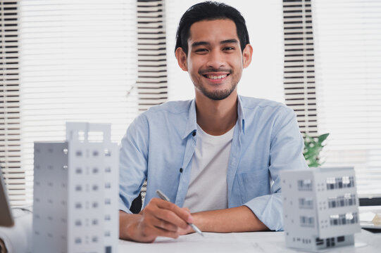 Smiling Asian architect working on building design project in modern office with architectural models. Business engineering, urban development, construction planning and real estate concept.