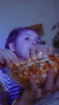 A girl watches something intently with her hand raised, seemingly asking for more. She holds a bowl of popcorn, indicating she is enjoying the moment.