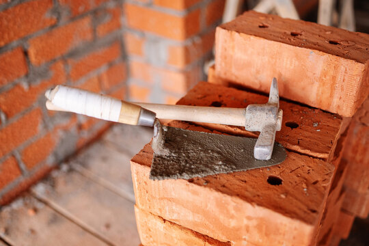 A dirty masonry trowel and a bricklayer's hammer rest on a stack of perforated red clay bricks against an unfinished brick wall at a construction site.