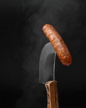 Grilled juicy sausage balanced on a sharp meat cleaver with steam on black background.