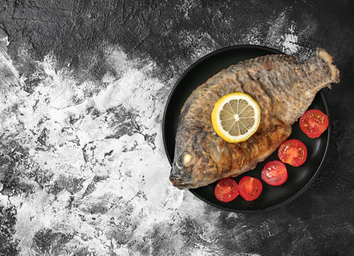 Roasted tilapia fish served with lemon slice and cherry tomatoes on a black plate.