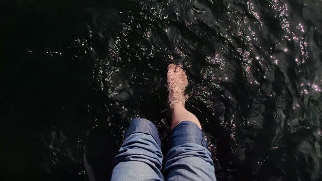 first person view adult man legs and bare feet dangling and kicking in cool, dark lake water on sunny day, creating ripples and enjoying moment summer freedom and relaxation.