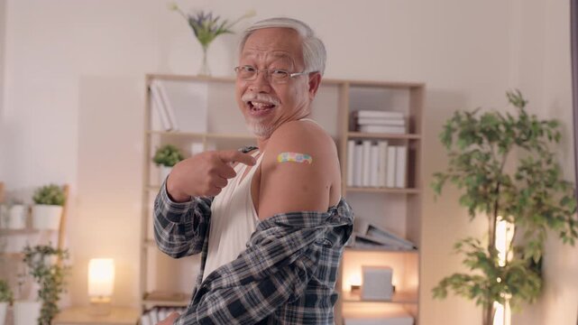 Vaccination senior man showing bandage upper arm after vaccination, smiling proudly home interior living room with eyeglasses and plaid shirt, happy healthcare prevention and immunity concept warm