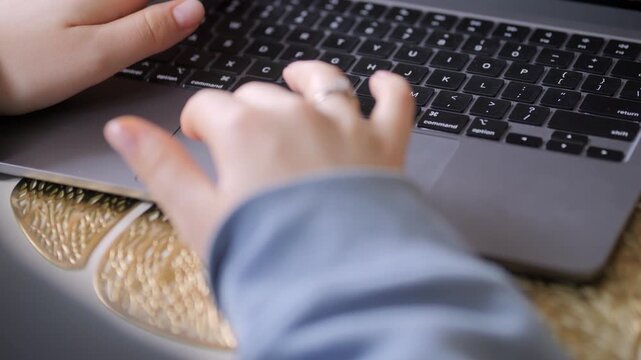 Hands typing on laptop keyboard. Wrist rests on woven mat. Fingers press keys. Person wears blue sleeve. Ring visible on finger. Laptop sits on textured surface. Close-up shot
