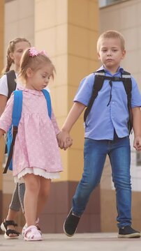 A girl in a pink dress walks beside a boy in a blue shirt. They are accompanied by an older girl, all carrying backpacks in an urban setting.