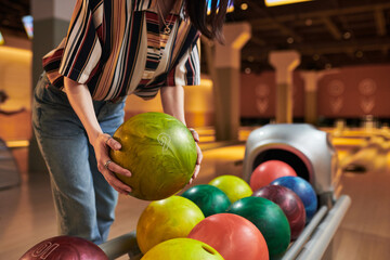 Fototapeta premium Young adult woman lifting bowling ball from rack in bowling alley, preparing recreational game and leisure activity. Useful for entertainment marketing, social outing promotion, close up