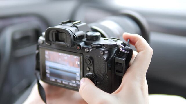 Hand holds camera inside car. Fingers adjust settings on back screen. Lens points forward, ready to capture. Interior details slightly blurred in background