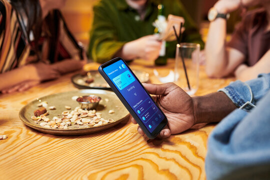 Close up of Black young adult man holding smartphone and sending digital payment while dining with friends, showing cashless transaction concept. Ideal for fintech, mobile banking, split bill